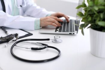 Close-up of stethoscope is lying on the table near female doctor typing on laptop computer